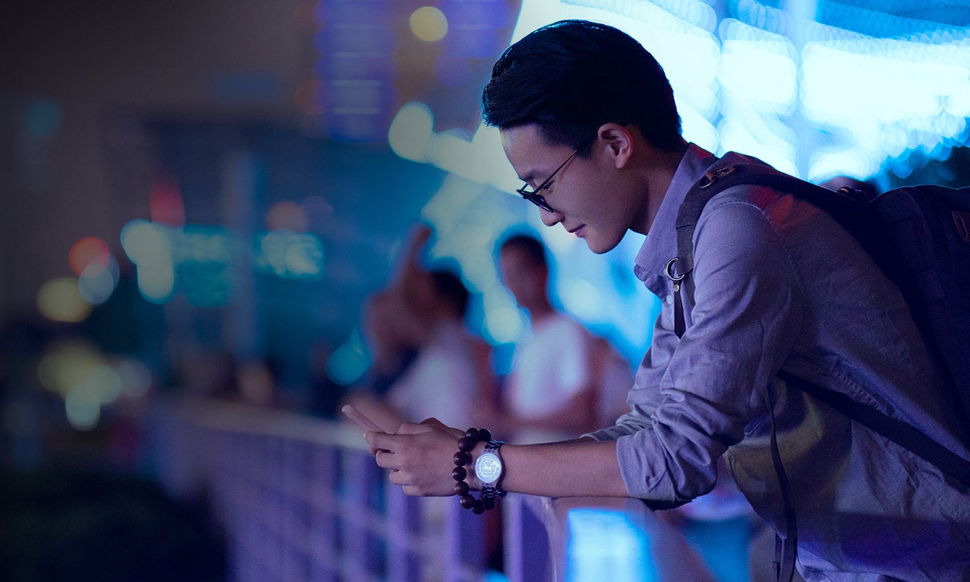 A young male in the foreground, leaning over a railing, looking at his cell phone